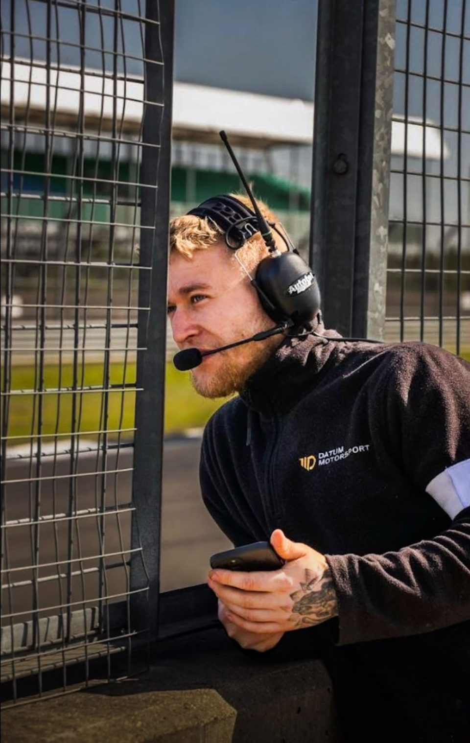 Sam Hayes at a circuit in team kit, wearing a radio headset and holding a timing screen.